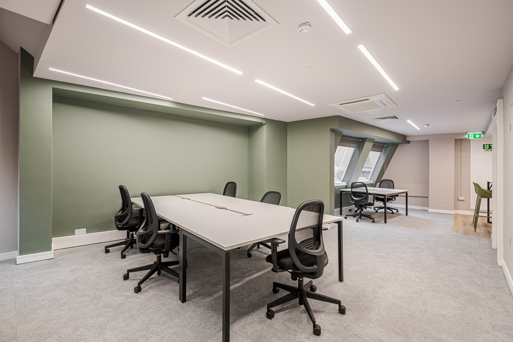 141 Fenchurch bright and airy office space designed for Alchemy Asset Management, featuring large white workstations, ergonomic black chairs, and a soft green accent wall. Recessed lighting and natural light from angled windows enhance the workspace.