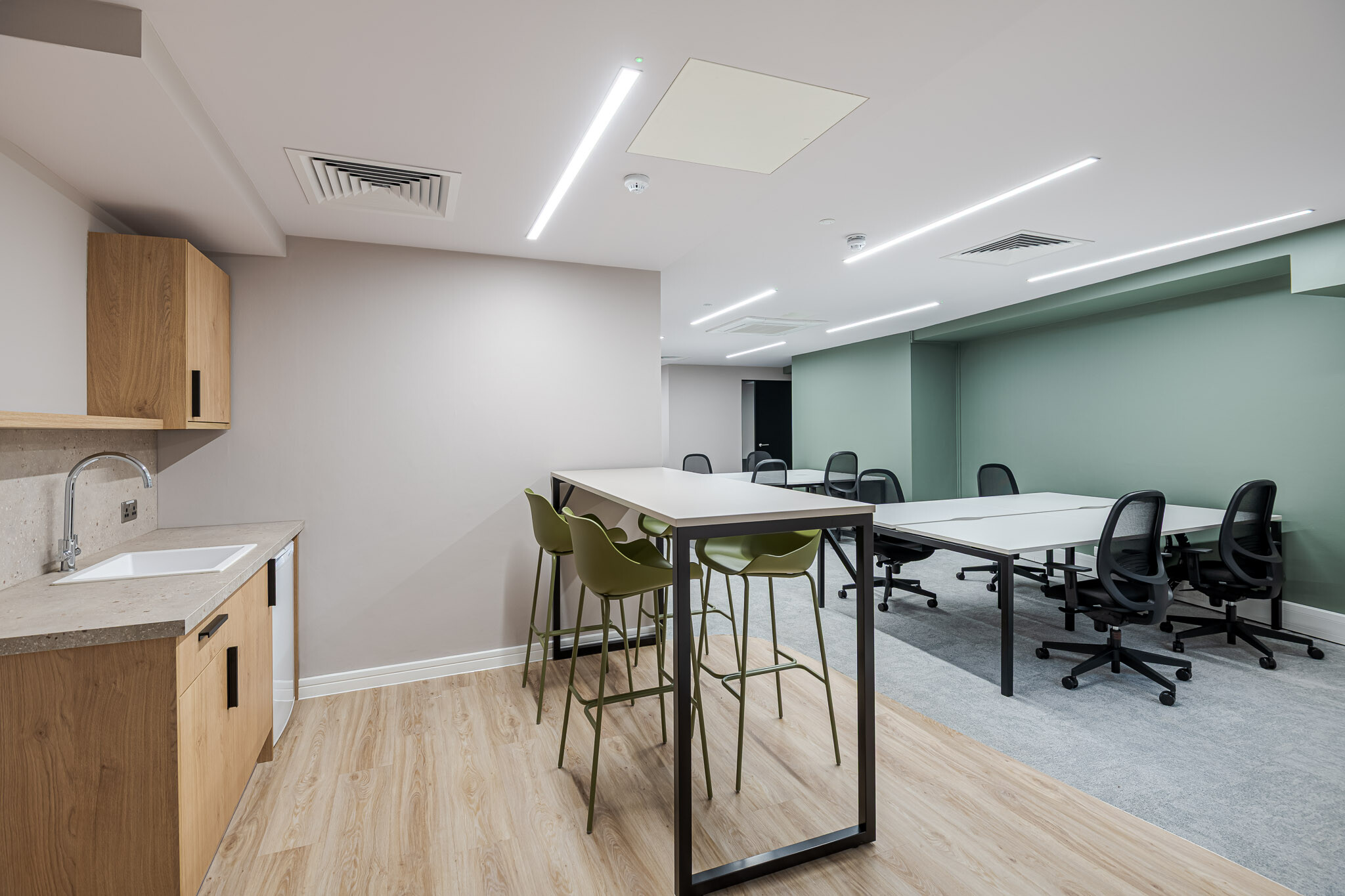 141 Fenchurch modern office space designed for Alchemy Asset Management, featuring a small teapoint with a wooden finish, a white sink, and a high table with olive green bar stools. In the background, workstations with ergonomic chairs and a muted green accent wall create a productive and stylish workspace.