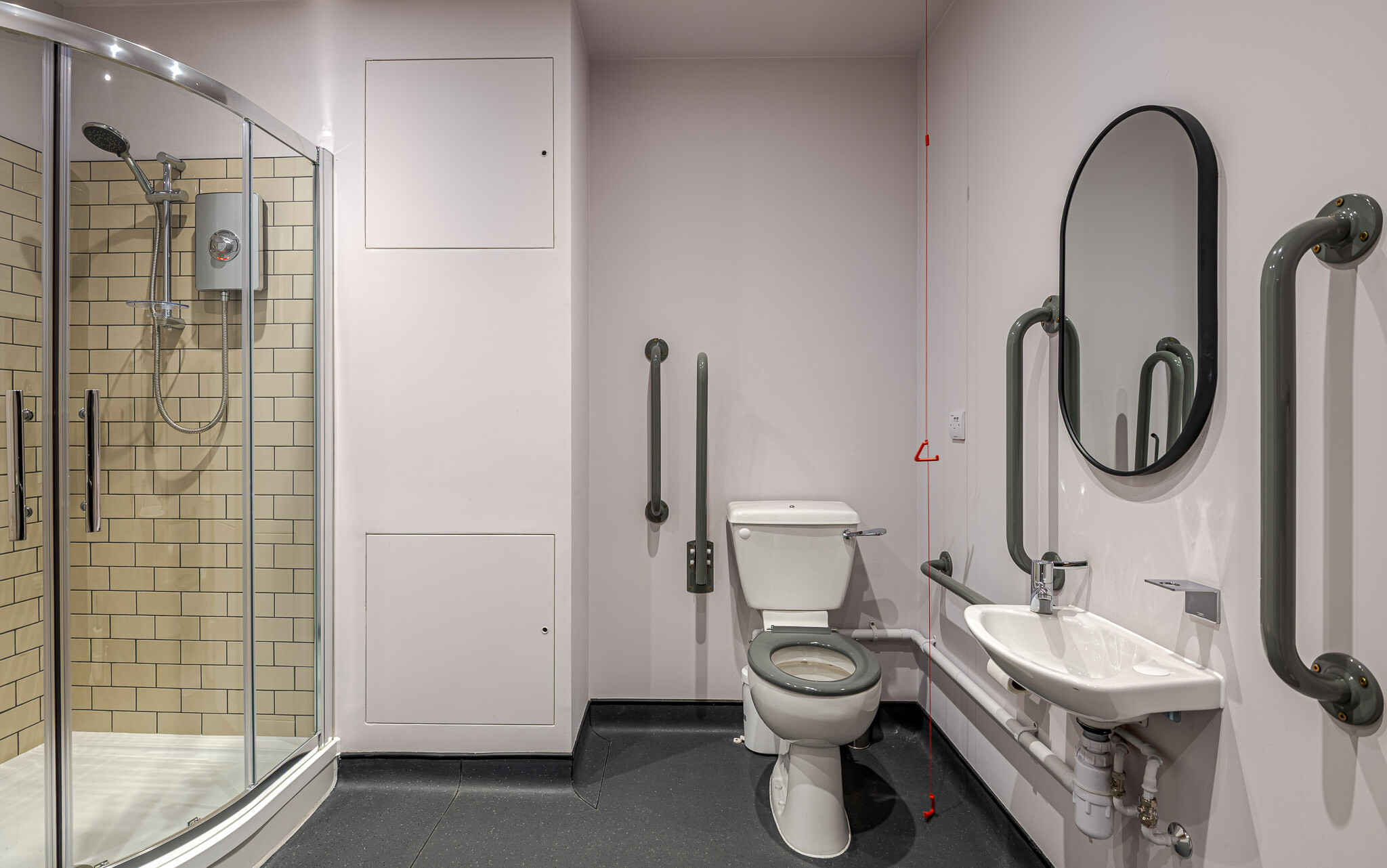 141 Fenchurch well-designed accessible bathroom for Alchemy Asset Management, featuring a walk-in shower with beige subway tiles, a toilet with grey grab rails, and a sink with an oval mirror. A red emergency pull cord ensures compliance with accessibility standards.