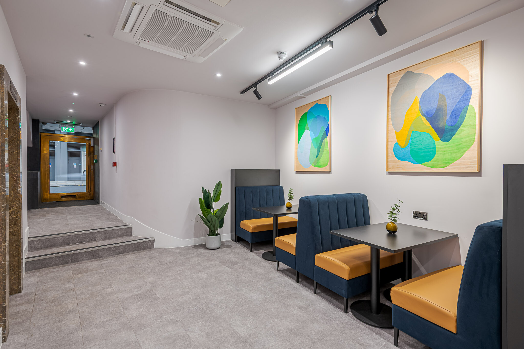 141 Fenchurch stylish breakout space created for Alchemy Asset Management, featuring navy and mustard booth seating, black tables, and vibrant abstract wall art. Soft lighting and indoor plants add warmth to the area, making it a great spot for informal meetings or relaxation.