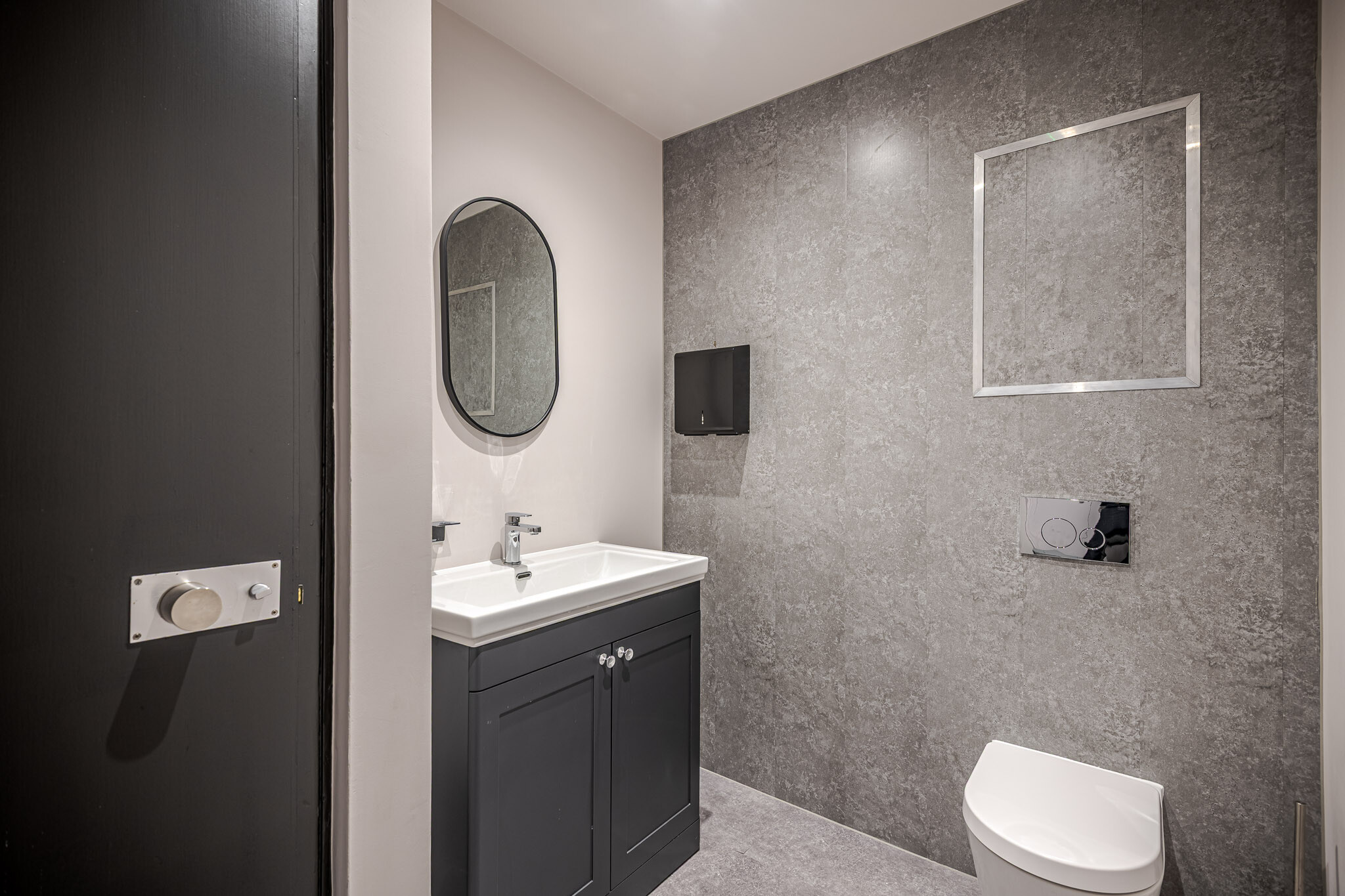141 Fenchurch sleek, modern bathroom designed for Alchemy Asset Management, featuring a dark vanity unit, a white sink, and a large oval mirror. The grey textured wall tiles add a sophisticated touch, while the minimalistic fixtures create a clean aesthetic.
