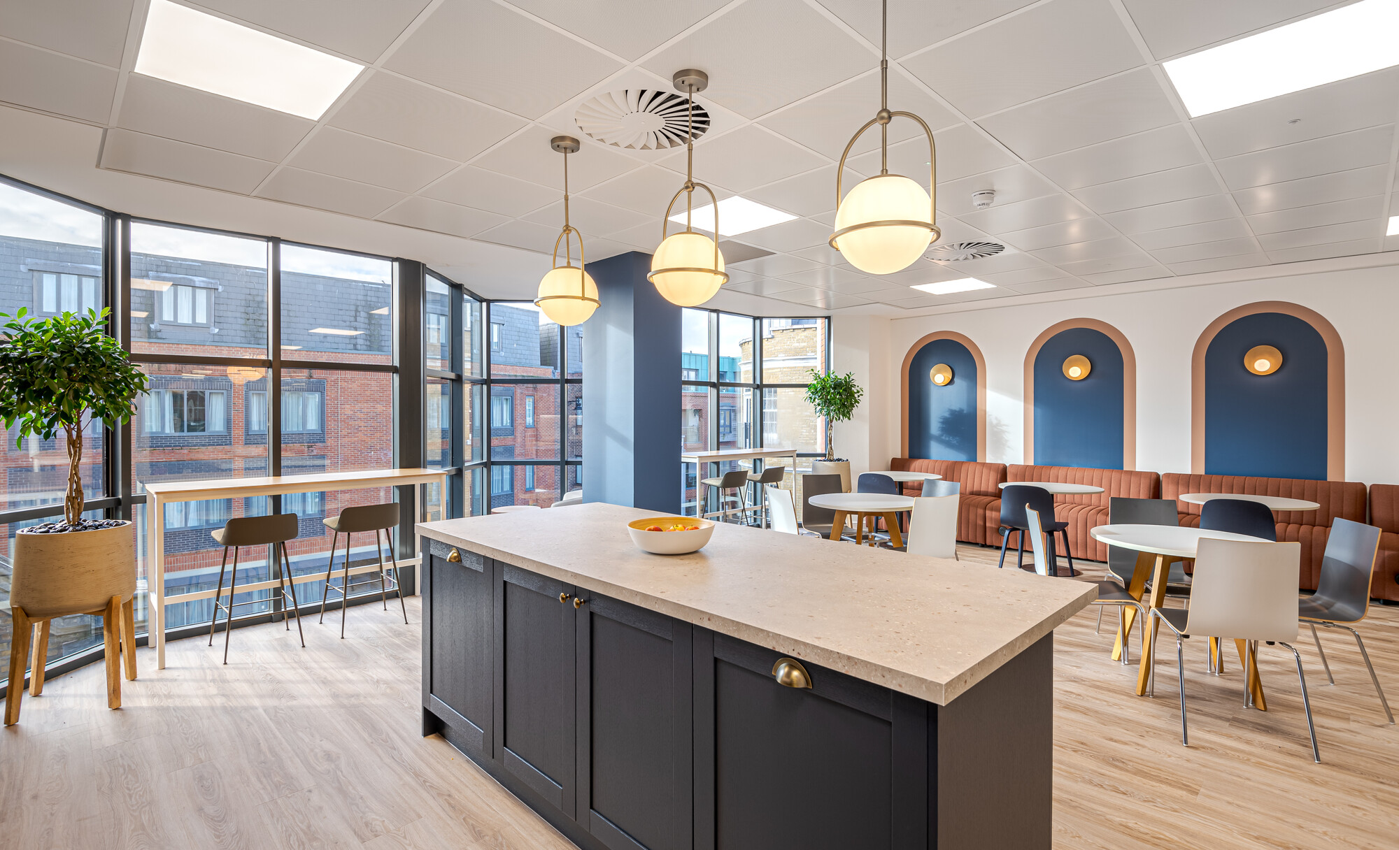 Accelya teapoint and breakout area with a central navy blue island and brass pendant lights. The space includes round tables, rust-coloured ribbed banquette seating, navy and white chairs, and large windows providing views of brick buildings. A tall potted plant and a high table with bar stools are also visible.