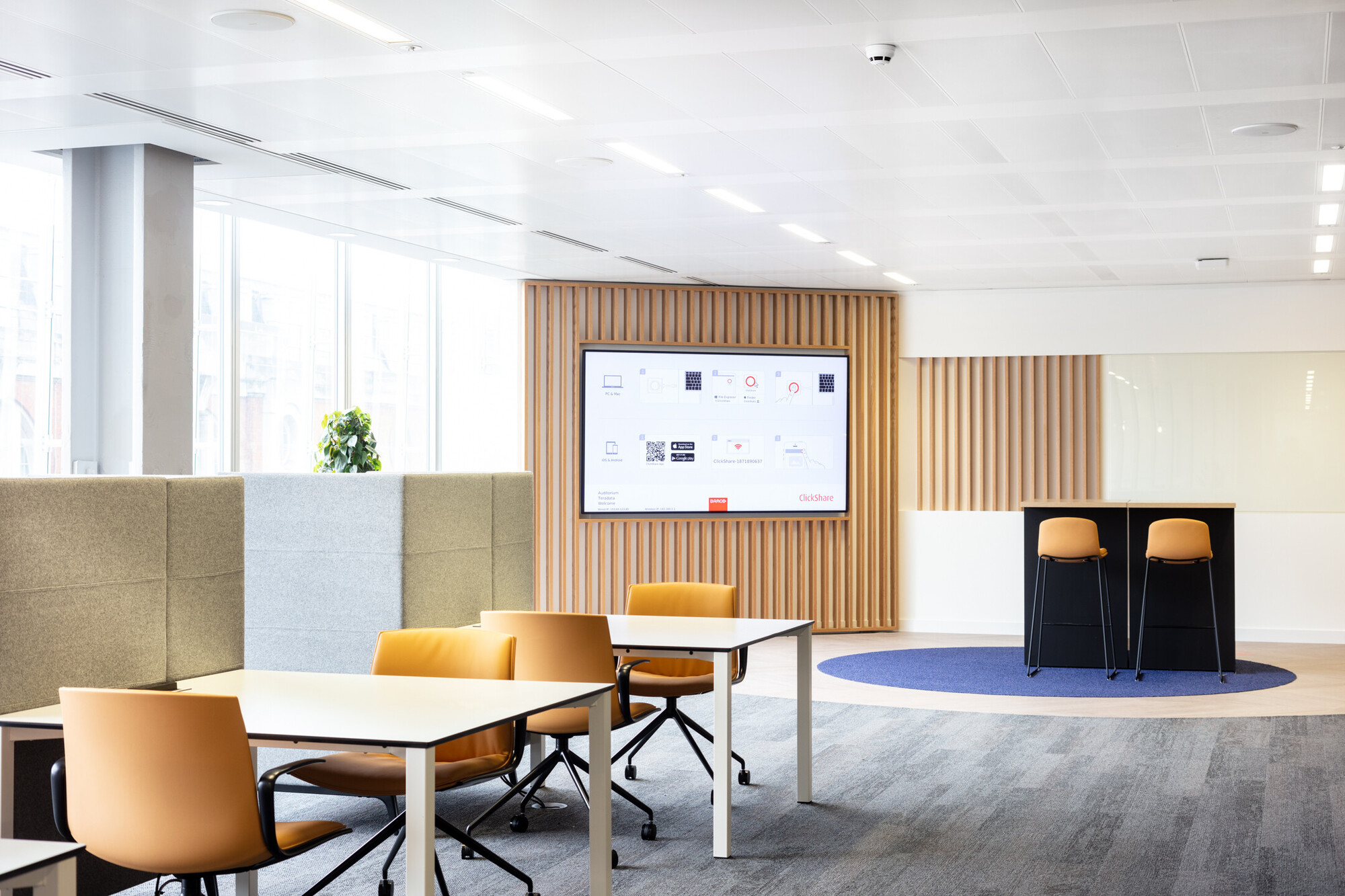 Contemporary Teradata office breakout area by Two, featuring a digital display board and casual seating with mustard chairs and round tables. The space includes high wooden dividers and soft carpet flooring for a cosy atmosphere.
