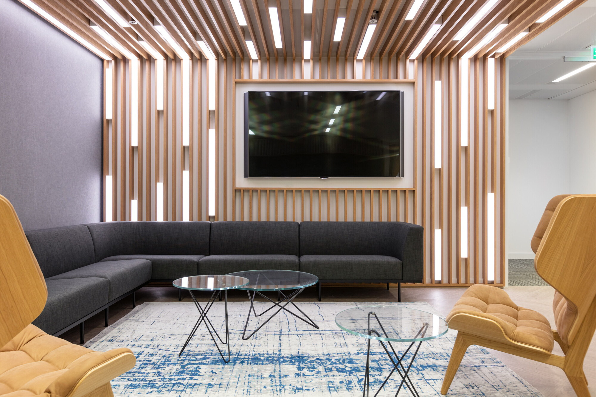 Chic Teradata office lounge area designed by Two, characterised by vertical wooden slats and recessed lighting. A grey sectional sofa pairs with modern wooden chairs and a glass coffee table over a vibrant blue area rug.