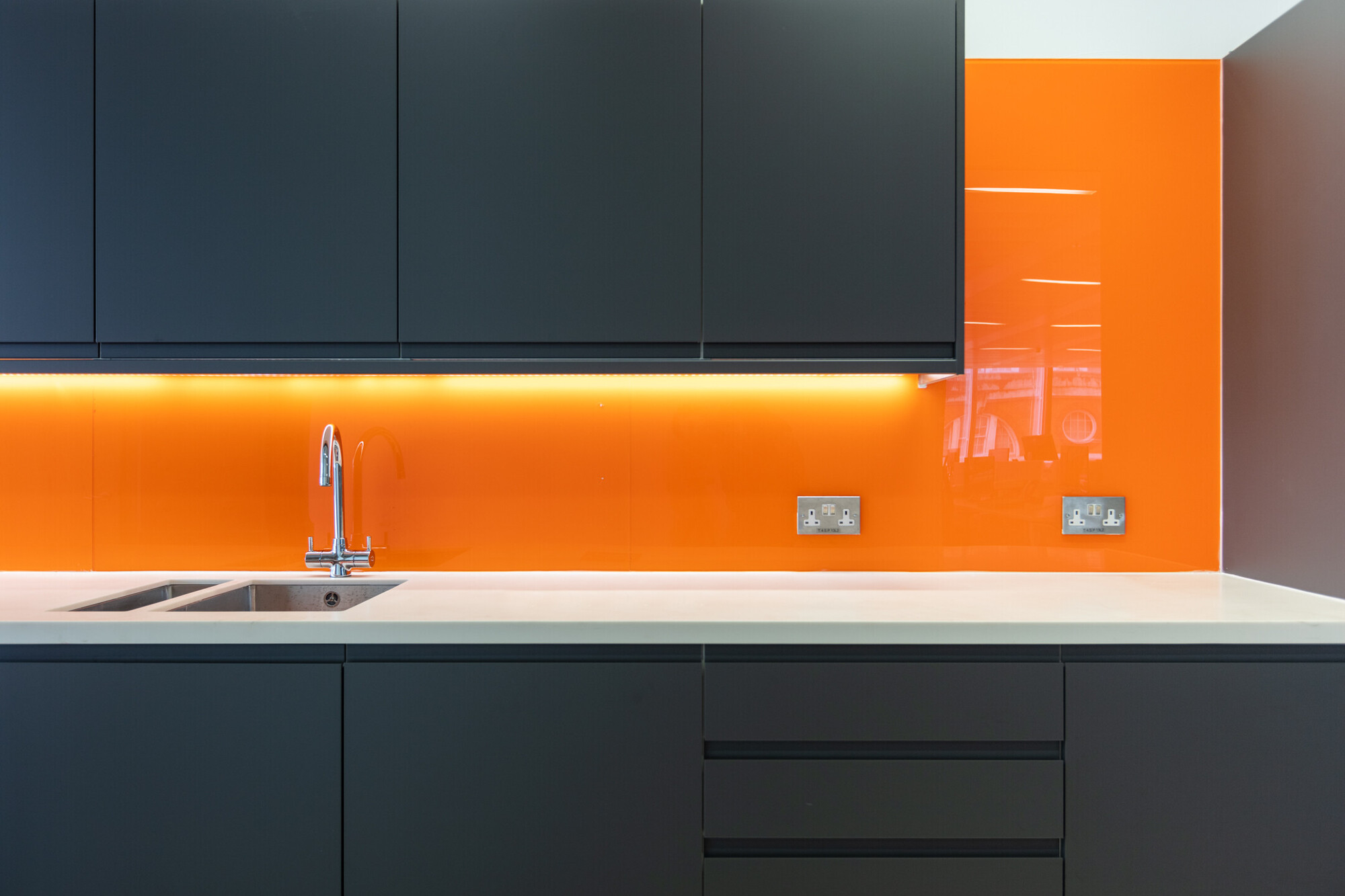 Bold and vibrant Teradata kitchen space in the office designed by Two, with striking orange backlighting and dark cabinetry. This modern kitchenette includes a white countertop and is equipped with high-end appliances