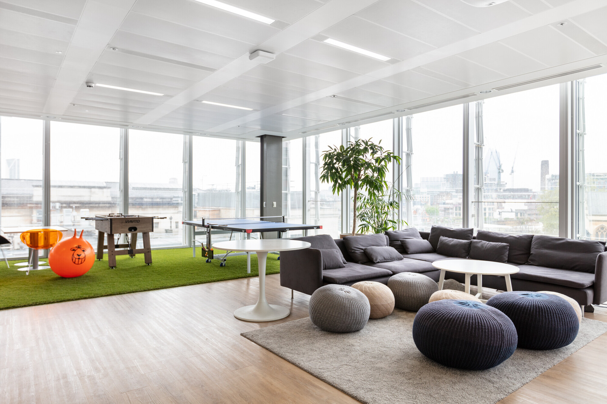 Spacious Teradata office lounge with floor-to-ceiling windows offering a panoramic city view, designed by Two. The area features a foosball table, ping pong table, and a relaxed seating arrangement with grey sofas and colourful poufs on a wooden floor.