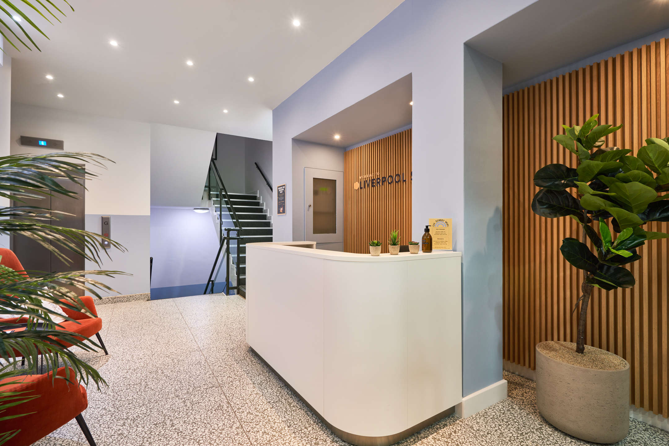 A stylish lounge at Work.life Liverpool Street by Two, featuring a deep blue sofa, artistic wall decor, and a view into contemporary workspace meeting rooms, ideal for collaboration and relaxation.